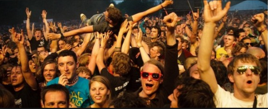 A large crowd of enthusiastic concertgoers is shown, with many people smiling, cheering, and raising their hands. One person is being lifted and crowd-surfing above the others. The atmosphere is vibrant and energetic, akin to the buzz created by successful social network marketing campaigns.