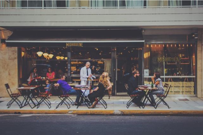 A bustling street-side café scene with several patrons seated at outdoor tables. The waiter, wearing a white apron, attends to a table while other diners engage in conversation. The café, named "MISHDIENE", features a modern storefront with glass windows and warm lighting, perfect for social media branding.