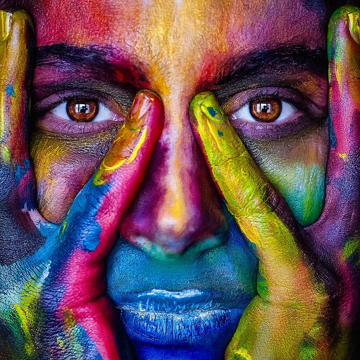 A close-up of a face covered in vibrant, multicolored paint. The person's hands, also painted in various shades, frame their eyes, which are looking directly at the camera. The striking colors create a dramatic and artistic effect, reminiscent of compelling content marketing visuals.