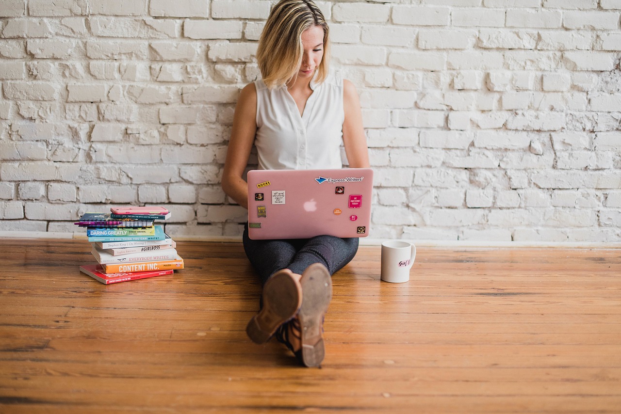 A person with shoulder-length blonde hair sits on a wooden floor against a white brick wall, working on a pink laptop adorned with stickers. Books are stacked to their left, and there's a white coffee mug nearby. Wearing a sleeveless white shirt and jeans, they focus intently on content marketing strategies.