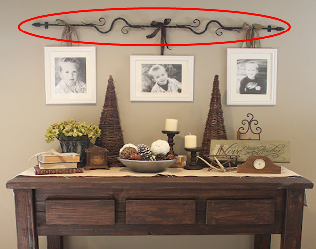 A wooden table with three framed photos on the wall above it, perfectly styled like a Single Pinterest Pin. Decorative items on the table include candles, a bowl of pine cones, a small clock, a vase with flowers, and cone-shaped tree decorations. An ornamental metal rod with a bow is mounted above the photos.
