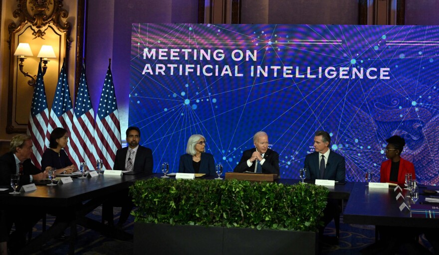 Seven people in business attire are seated at long tables arranged in a U-shape at a conference. A sign behind them reads "Meeting on Artificial Intelligence." American flags are placed in the room. A man in the middle is speaking about AI, while others listen attentively.